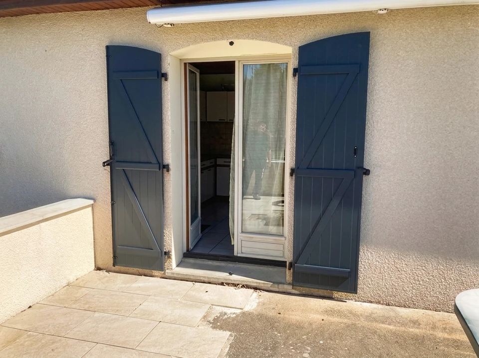 Façade extérieure de maison avec deux fenêtres à volets bleus ouverts, mur en brique blanche sous porche couvert