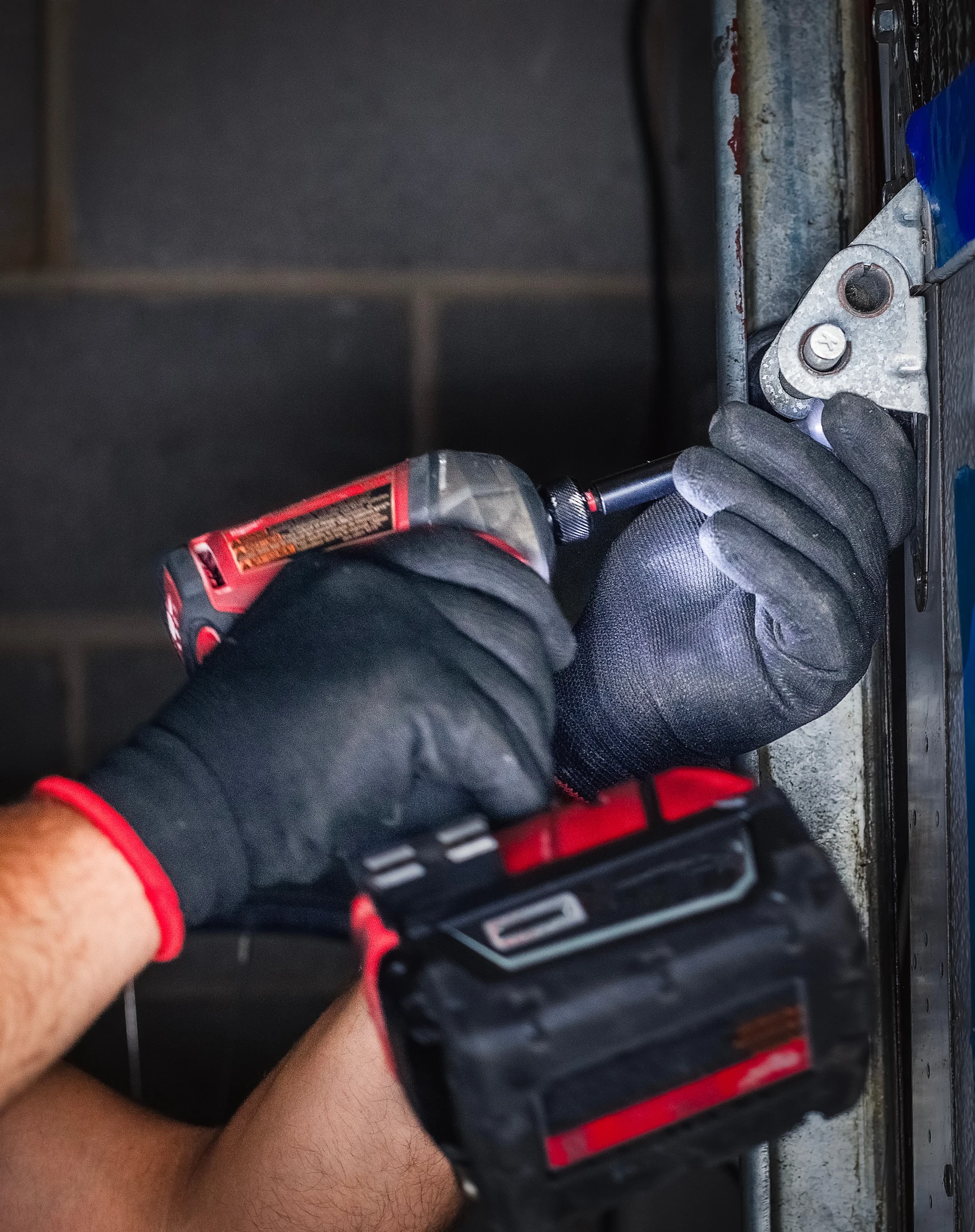 Un artisan menuisier en tenue de travail professionnel installant une porte de garage, effectuant des réglages ou des tests de fonctionnement, montrant son expertise technique lors de la pose. La porte de garage est clairement visible et en cours de travaux ou nouvellement installée. La scène se déroule en extérieur ou semi-extérieur devant un garage de maison résidentielle ou professionnelle.