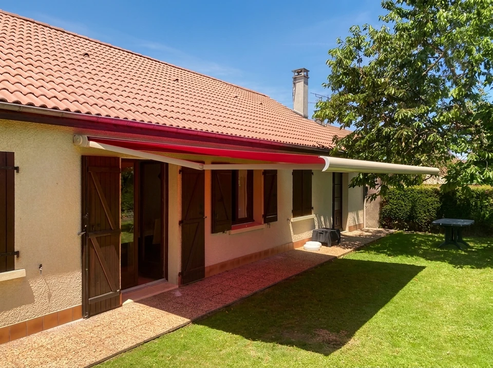 Gros plan sur la facade d'une maison moderne avec une grande baie vitrée exposée au soleil du midi. Un store brise-soleil blanc ou gris perle est partiellement déployé, créant une zone d'ombre nette sur la vitre et la terrasse devant. Le contraste entre la zone ensoleillée brillante et la zone à l'ombre du store est clairement visible. On voit par la vitre l'intérieur frais et lumineux de la maison. Les stores doivent être de type store enrouleur, banne ou extérieur, professionnels et modernes.